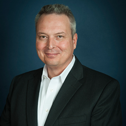 Man in a dark suit and white shirt, smiling against a dark blue background.