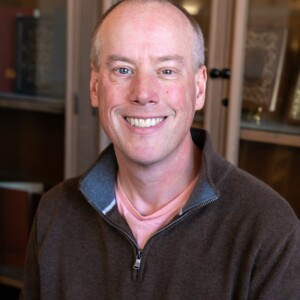 Smiling person with short hair in a brown sweater; bookshelves in the background.