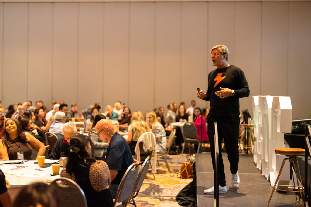 conference-speaker-addresses-audience-1.jpg Speaker addresses conference audience from stage; attendees seated at tables, listening.